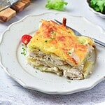 A slice of baked chicken lasagna with a golden, melted cheese topping is served on a white plate. A fork rests beside the lasagna, and a halved cherry tomato and herbs garnish the plate.