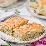 Two rectangular pieces of golden-brown, dense homemade cake are placed on a decorative white plate, with a fork and another plate of cake visible in the blurred background, evoking comfort much like a classic Corn Casserole recipe.