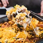 A close-up of a hand holding a spatula lifting a serving of baked casserole with melted cheese from a square baking pan. The casserole contains pasta, vegetables, and a golden topping.