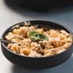 A black bowl filled with creamy pasta mixed with sauce and herbs, garnished with a fresh basil leaf, features a delicious Slow Cooker chicken recipe inside, with a spoon resting in the bowl.
