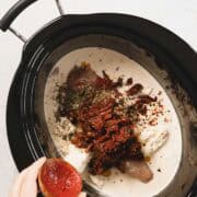 A hand holds half a tomato over a slow cooker filled with raw chicken breasts, cream, dried herbs, sun-dried tomatoes, and red pepper flakes—perfect for a Creamy Sun Dried Tomato Chicken recipe.