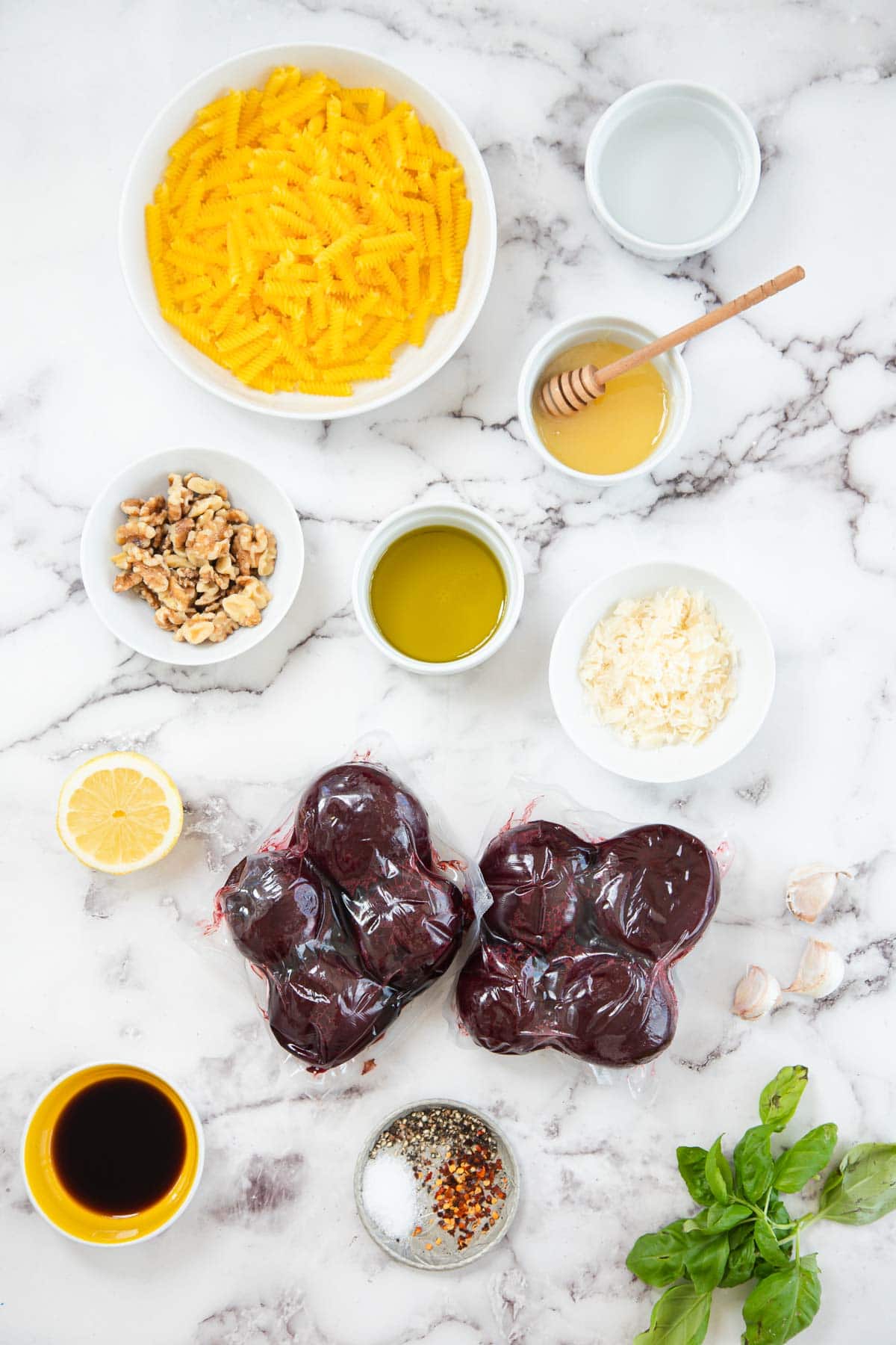 A flat lay of raw chicken livers, dried pasta, walnuts, grated parmesan cheese, fresh basil, a lemon half, garlic cloves, olive oil, honey, soy sauce, seasonings, and a small bowl of water&mdash;perfect for a unique walnut pasta sauce.