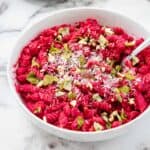 A white bowl filled with rotini pasta coated in a vibrant beet pasta sauce, topped with grated cheese, chopped nuts, and fresh basil leaves, set on a marble surface. A fork rests in the bowl.
