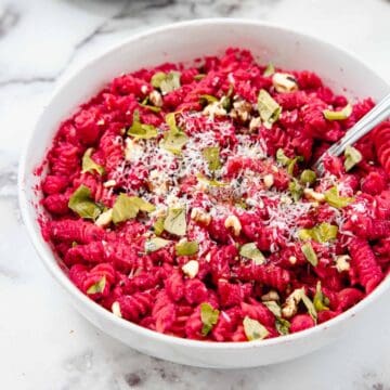 A white bowl filled with rotini pasta coated in a vibrant beet pasta sauce, topped with grated cheese, chopped nuts, and fresh basil leaves, set on a marble surface. A fork rests in the bowl.