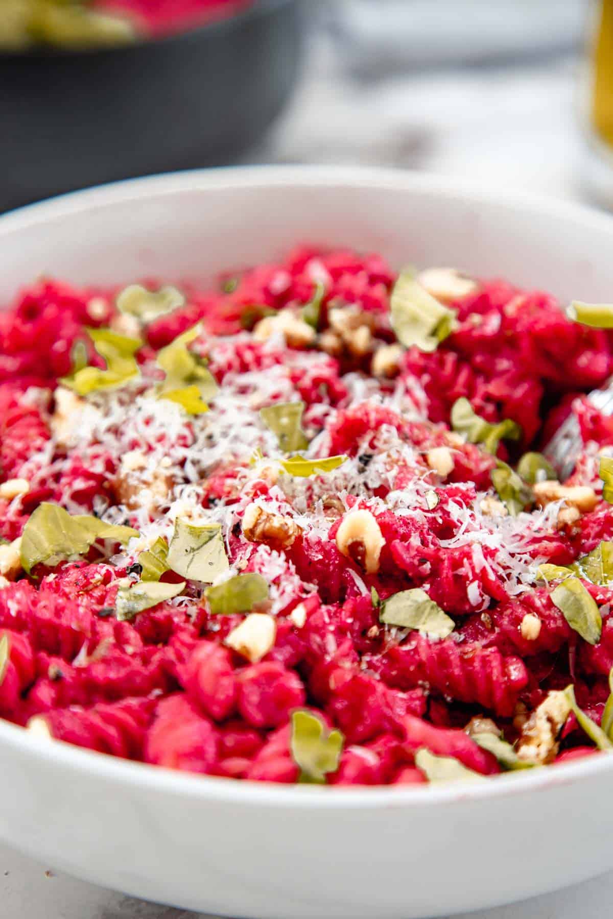 A white bowl filled with bright red risotto, likely made with beet pasta sauce, topped with grated cheese, chopped walnuts, and fresh green herbs. The background is blurred with hints of another bowl or pan.
