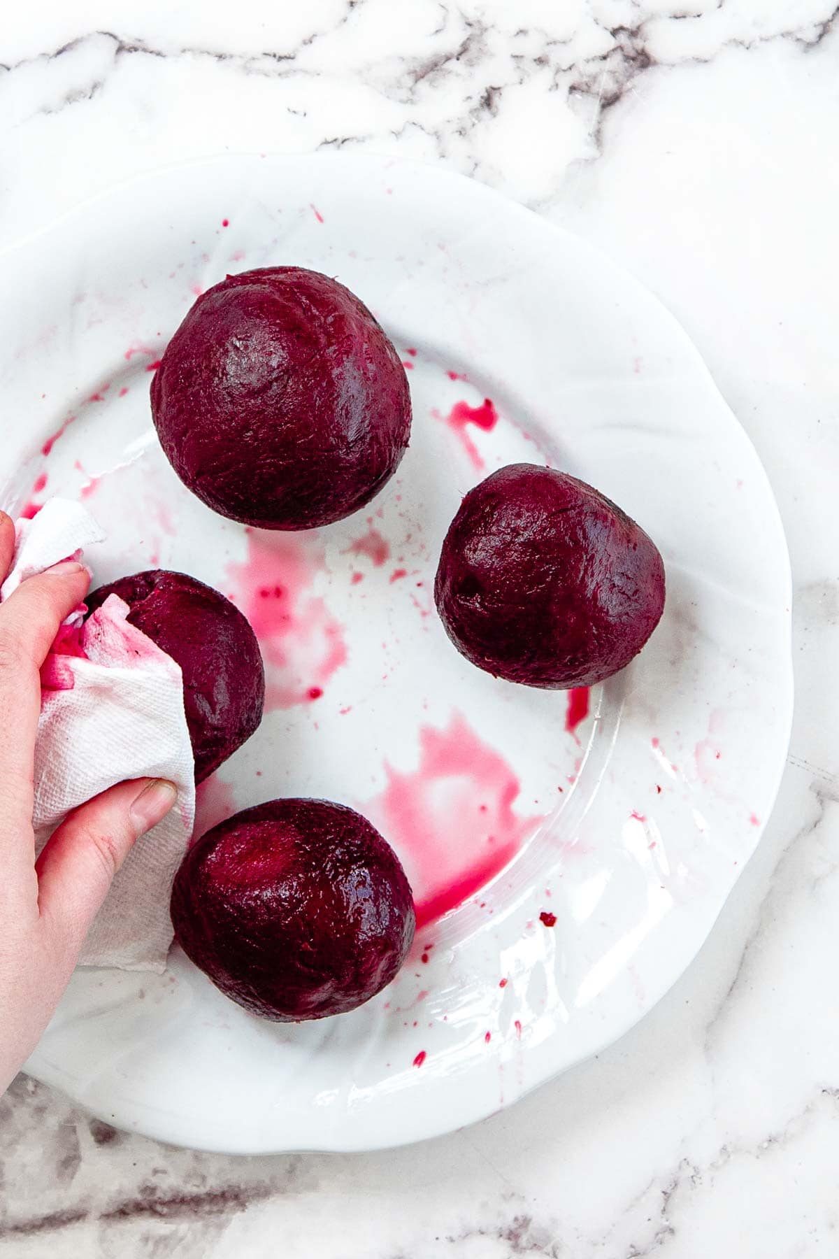 A hand uses a paper towel to wipe beet juice from a white plate holding four whole, peeled beets&mdash;perfect for making beet pasta sauce. The plate sits on a marble surface.