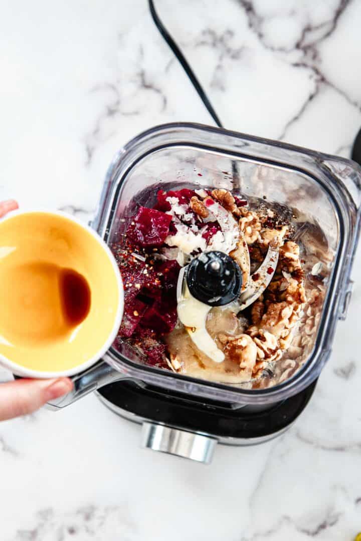 A blender filled with beetroot, walnuts, onion, and spices sits on a counter. A hand pours a brown liquid into the mix, ready to create a vibrant Blender Beet Pasta Sauce. The background is a white, marbled surface.