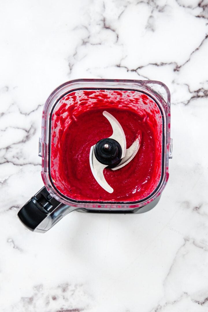 A food processor with a sharp metal blade is filled with a thick, bright red beet pasta sauce. The appliance sits on a white marble surface with elegant gray veins.