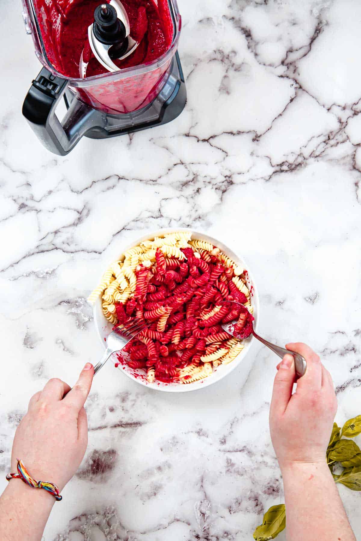 A person mixing cooked rotini pasta with bright red Blender Beet Pasta Sauce in a white bowl on a marble countertop. A food processor with sauce inside is nearby, along with a bunch of leafy greens and some walnuts partially visible.
