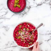 A white bowl of pink pasta with herbs, grated cheese, and Blender Beet Pasta Sauce sits with a fork inside and a hand holding the bowl. Above is more vibrant pasta sauce garnished with basil leaves on a white marble surface.