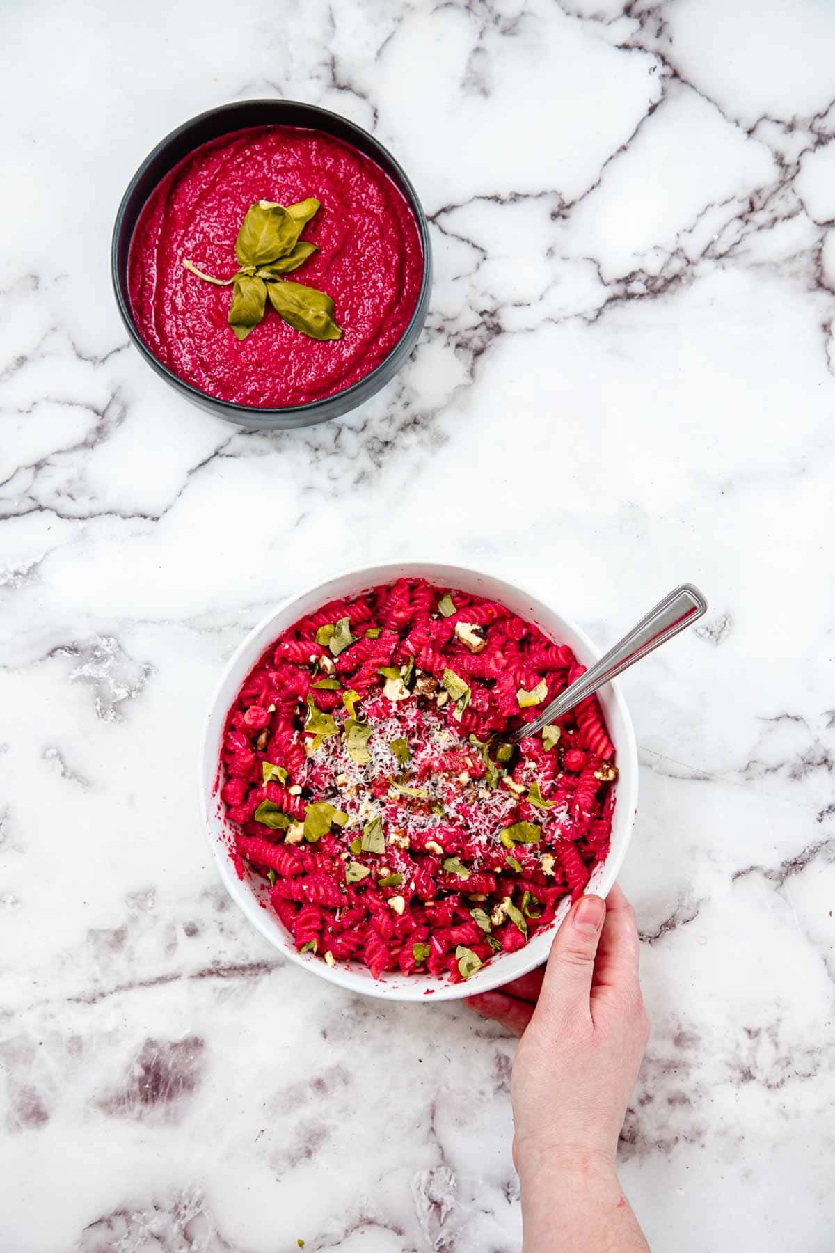A white bowl of pink pasta with herbs, grated cheese, and Blender Beet Pasta Sauce sits with a fork inside and a hand holding the bowl. Above is more vibrant pasta sauce garnished with basil leaves on a white marble surface.