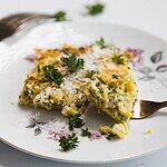 A close-up of a slice of herb and cheese frittata on a white floral plate, resembling a crustless zucchini quiche, garnished with grated cheese and parsley, with a fork holding a bite-sized piece.