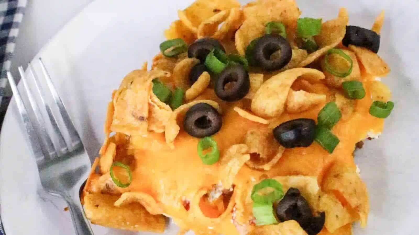 A plate with a serving of cheesy casserole topped with corn chips, black olives, and green onions. A portion is served on a white plate with a fork, next to a checkered cloth.