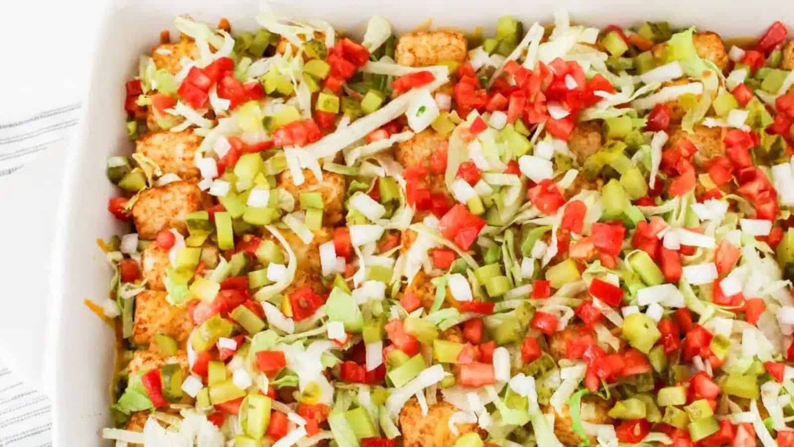 A casserole dish filled with tater tots, ground beef, diced tomatoes, shredded lettuce, onions, and pickles, with a serving spoon on the side.