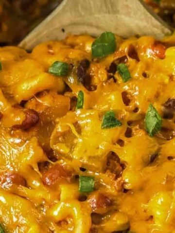 Close-up of cheesy macaroni mixed with vegetables and beans, topped with melted cheddar cheese and garnished with chopped green onions. A wooden spoon is partially visible in the background.