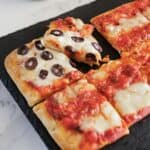 Rectangular pieces of flatbread pizza on a black tray, some topped with tomato sauce and melted cheese, and one slice with black olive slices being pulled away—an easy recipe perfect for your 4th of July gathering.