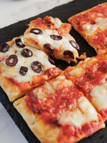 Rectangular pieces of flatbread pizza on a black tray, some topped with tomato sauce and melted cheese, and one slice with black olive slices being pulled away—an easy recipe perfect for your 4th of July gathering.
