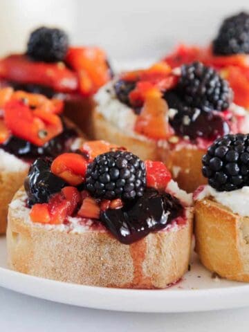 A plate of sliced baguette topped with a layer of white cheese, mixed berry jam, chopped red fruit, and fresh blackberries, arranged on a white plate on a light surface.