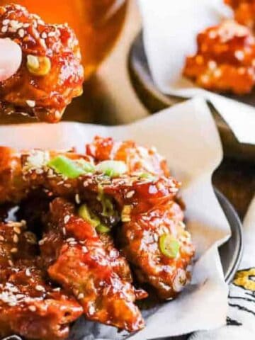 A hand holding a piece of saucy fried chicken garnished with sesame seeds and green onions, with more pieces on a plate lined with parchment paper nearby.