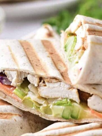 Two grilled wraps are cut in half, showing fillings of grilled chicken, lettuce, cucumber, and other vegetables. The wraps have visible grill marks and are stacked on a plate with a blurred background.