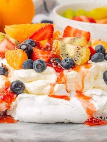 A pavlova dessert topped with whipped cream, sliced strawberries, blueberries, kiwi, and orange segments sits on a marble surface, with bowls of fruit and whole oranges in the background.
