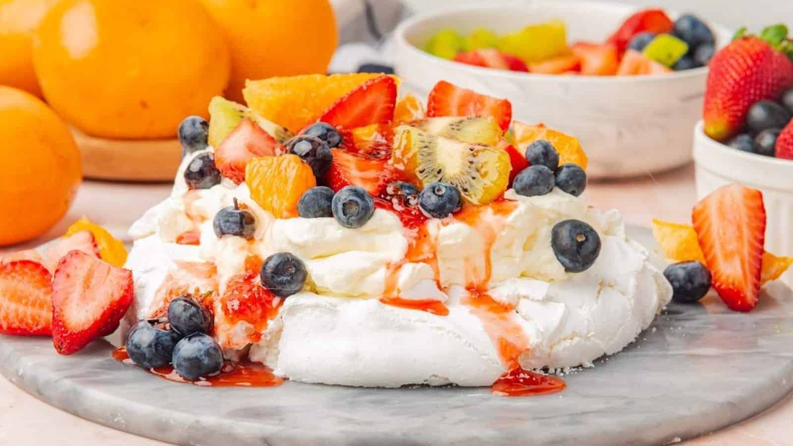 A pavlova topped with whipped cream, strawberries, blueberries, kiwi, and orange slices on a marble surface, surrounded by whole strawberries and oranges.