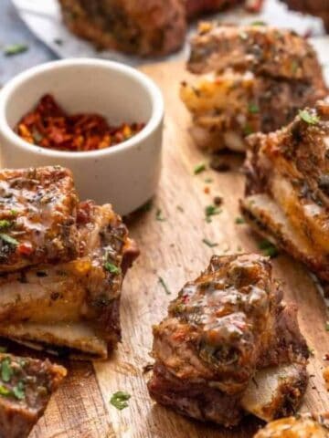Several pieces of seasoned, cooked beef ribs are arranged on a wooden cutting board, garnished with chopped herbs. Small bowls with spices and sauce are in the background.
