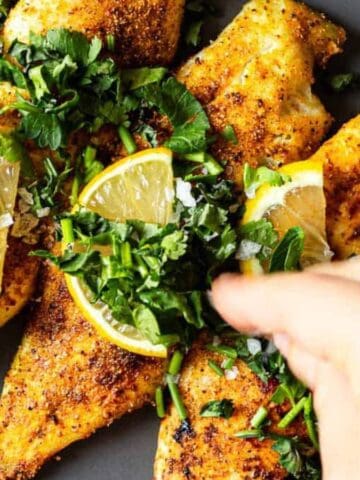 A hand sprinkles chopped herbs over cooked, seasoned chicken breasts garnished with lemon slices, all arranged on a gray plate.