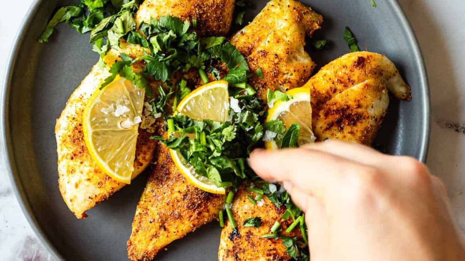 A hand sprinkles chopped herbs over cooked, seasoned chicken breasts garnished with lemon slices, all arranged on a gray plate.