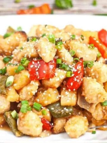 A square white plate holds a serving of sweet and sour chicken with red and green bell peppers, pineapple chunks, and chopped green onions, garnished with sesame seeds. A fork rests on the side of the plate.