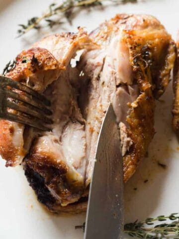 A close-up of a cooked chicken thigh being cut with a knife and held with a fork on a white plate, garnished with fresh thyme sprigs. The chicken has crispy, browned skin.