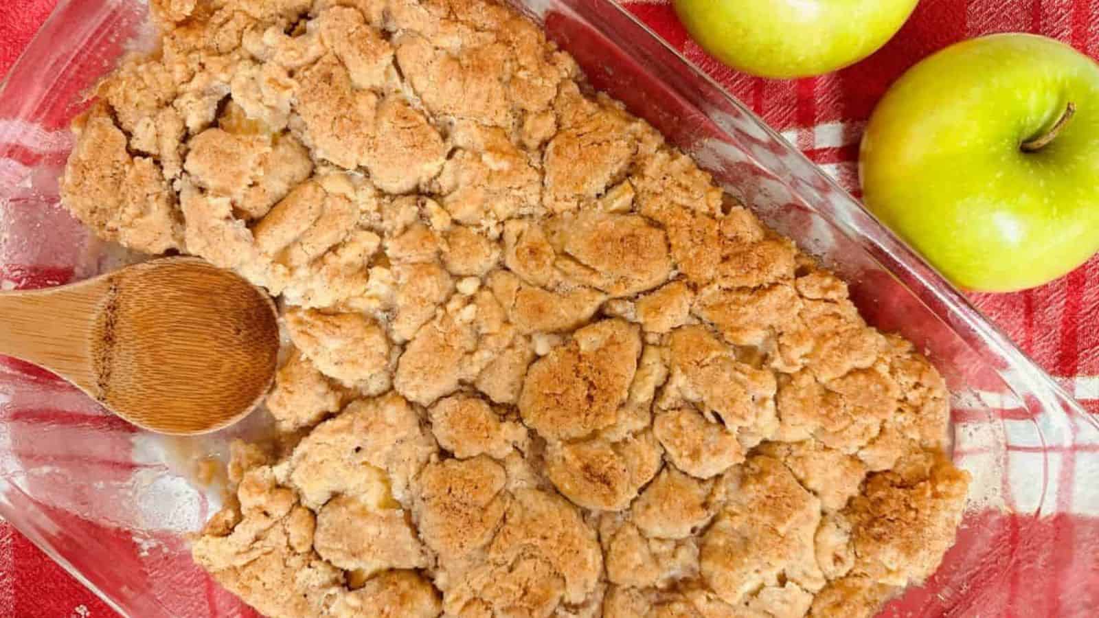 Apple crisp in a glass dish with apples and a wooden spoon.