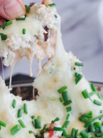 A hand lifts a portion of cheesy dip topped with chopped chives from a dish, with melted cheese stretching and blue tortilla chips visible in the background.