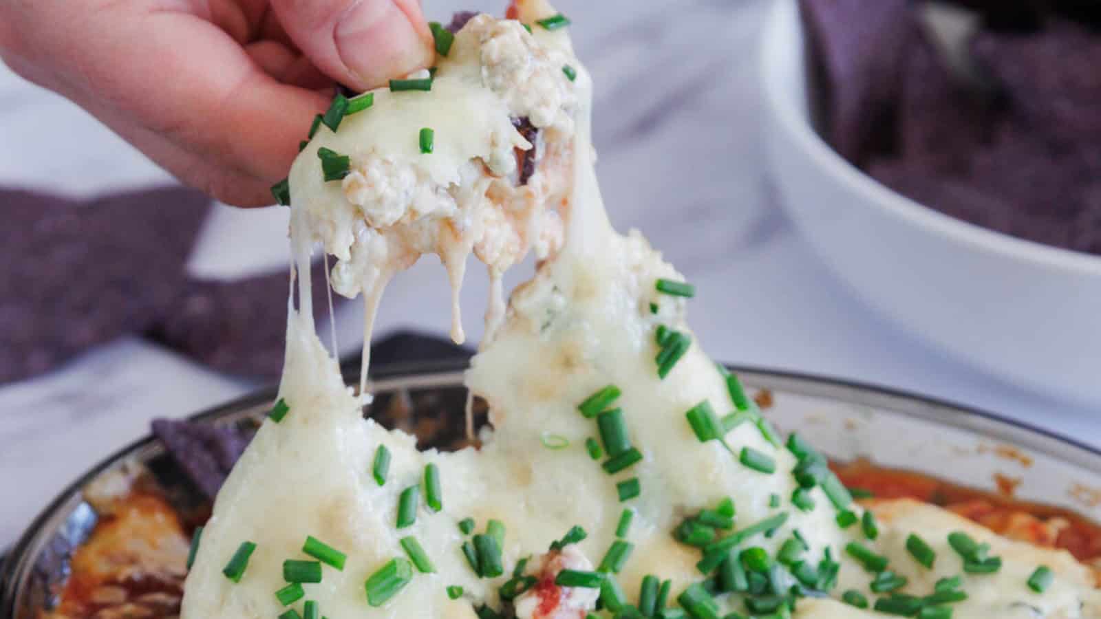 A hand lifts a portion of cheesy dip topped with chopped chives from a dish, with melted cheese stretching and blue tortilla chips visible in the background.