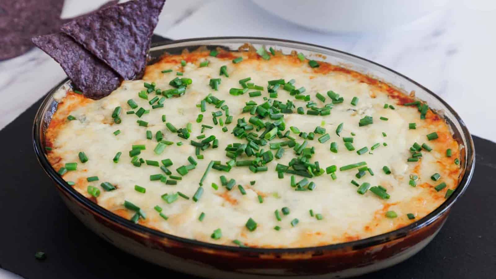 A round glass dish filled with a baked cheesy casserole topped with chopped green chives. Two triangular blue corn tortilla chips are inserted upright on one side of the dish.
