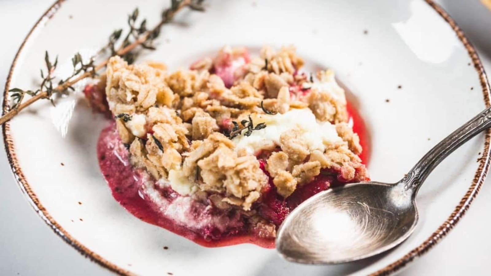 A serving of fruit crumble with a crumbly oat topping and a layer of red fruit filling, garnished with a sprig of thyme on a white plate with a brown rim, next to a silver spoon.