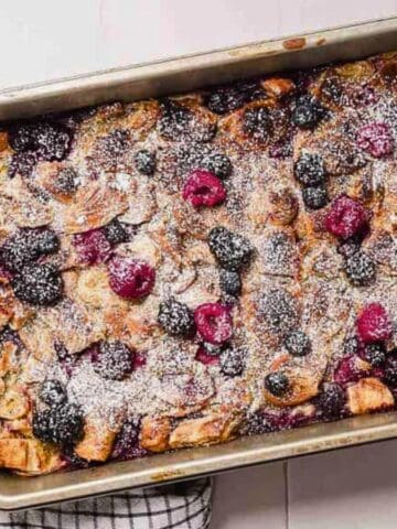 A rectangular metal baking pan filled with baked bread pudding topped with mixed berries and dusted with powdered sugar, sitting on a white tiled surface next to a black and white checkered towel.
