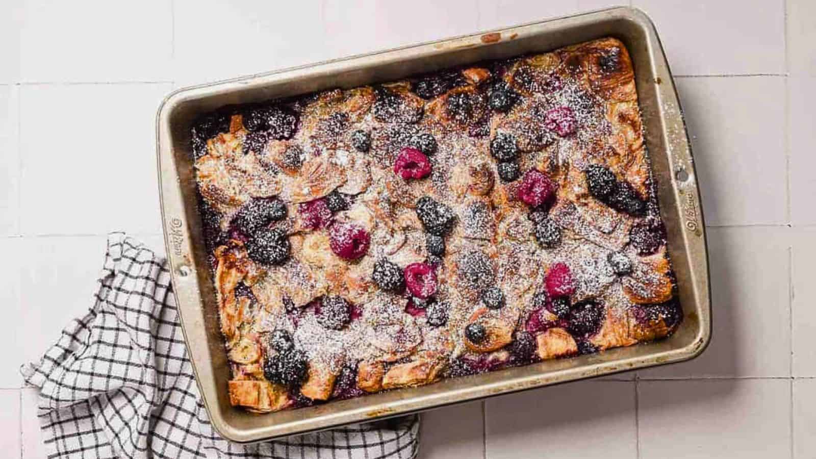 Close-up of a baked dessert topped with powdered sugar, featuring blackberries, raspberries, and blueberries. The dish is in a rectangular baking pan, with visible golden-brown edges and drizzles of cream or sauce.