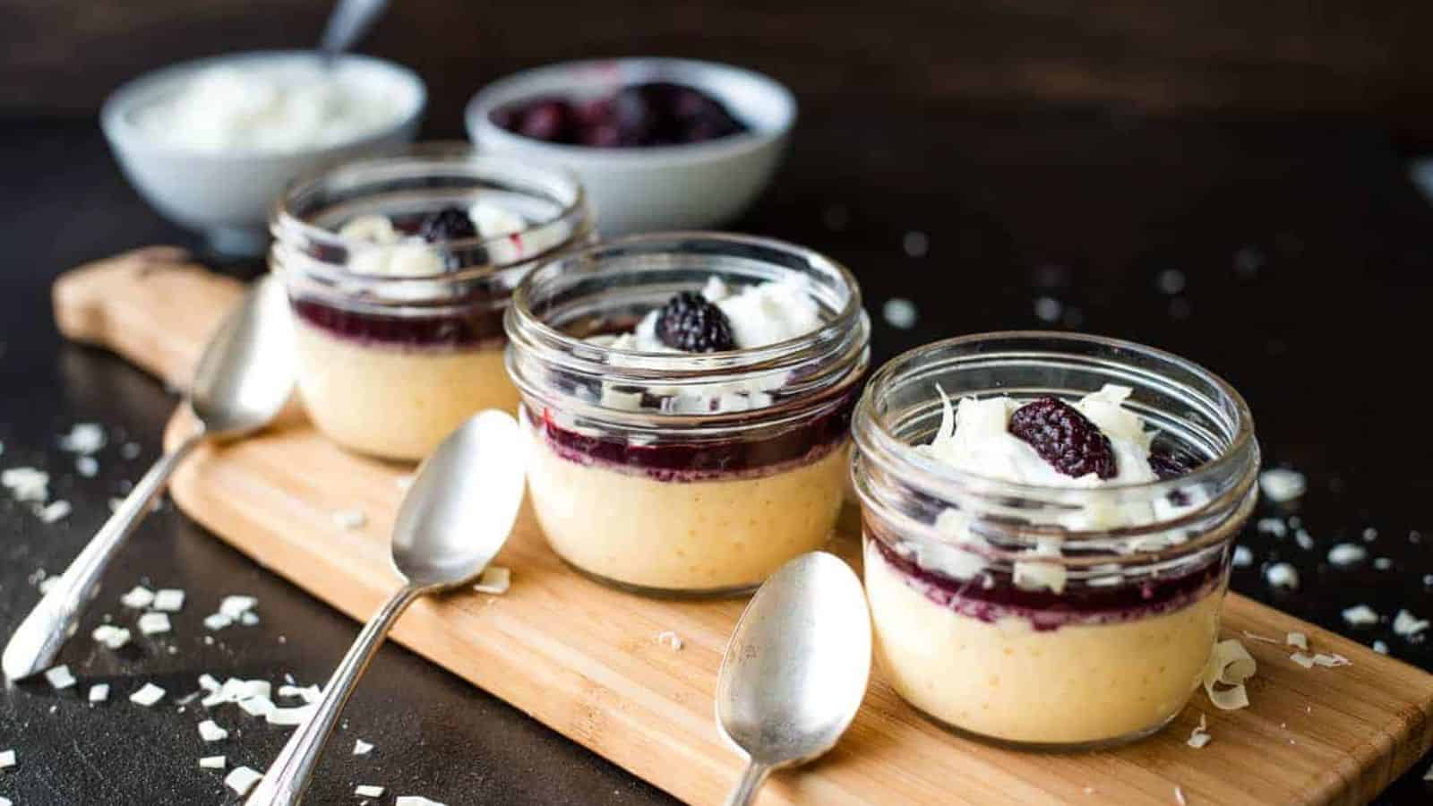 Three blackberry pots de creme lined up on a wooden board next to silver spoons.