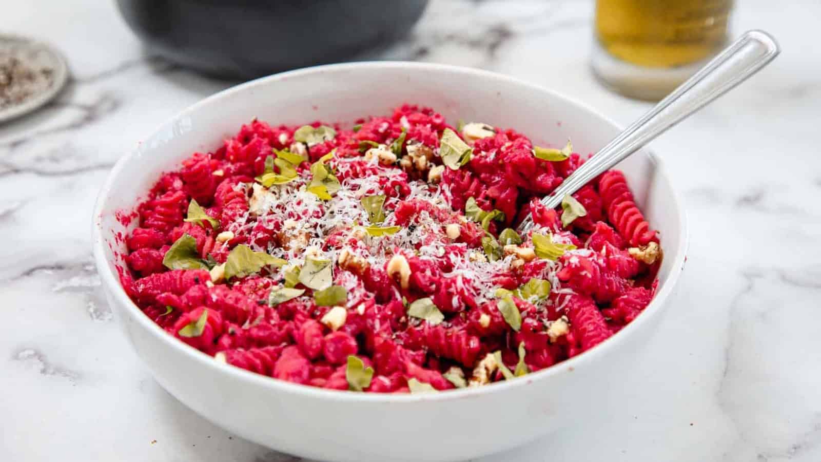 A white bowl filled with rotini pasta coated in bright pink beet sauce, topped with grated cheese, chopped nuts, and fresh herbs, with a spoon resting in the bowl on a marble surface.