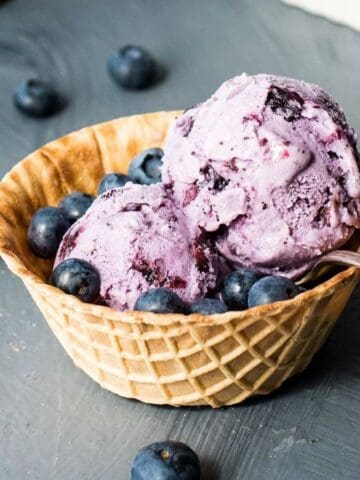 A waffle bowl filled with two scoops of blueberry ice cream, garnished with fresh blueberries, sits on a gray surface with additional blueberries scattered around. A metal spoon rests in the bowl.