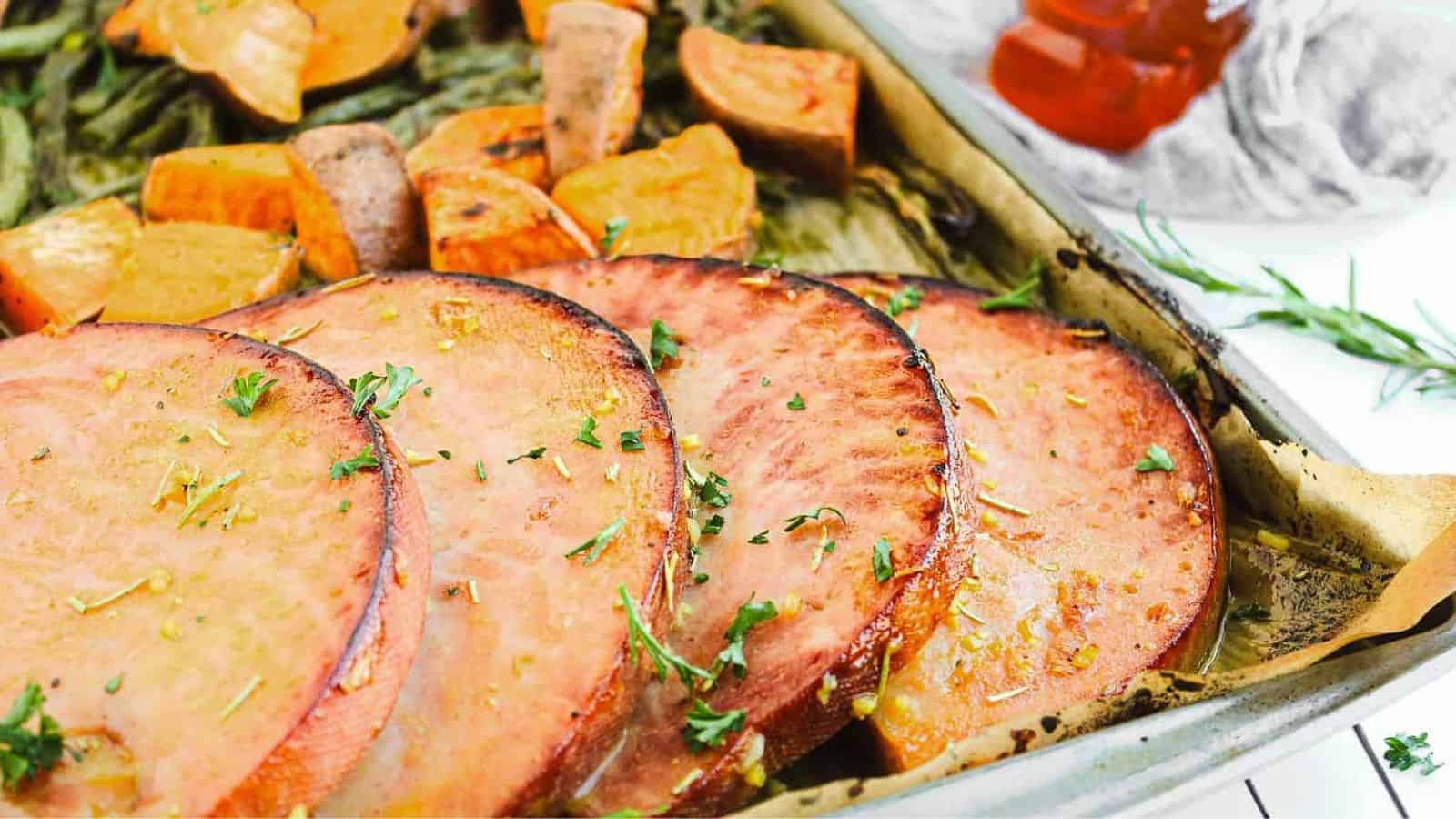 A white plate with a serving of glazed ham garnished with rosemary, surrounded by roasted sweet potato chunks and green beans. A gray napkin and a bottle of syrup are beside the plate.