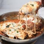 A close-up of baked eggplant parmesan in a metal pan. A spatula lifts a cheesy, sauce-covered serving with melted mozzarella stretching from the pan. Fresh basil pieces are visible on top.