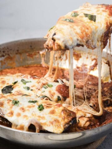 A close-up of baked eggplant parmesan in a metal pan. A spatula lifts a cheesy, sauce-covered serving with melted mozzarella stretching from the pan. Fresh basil pieces are visible on top.