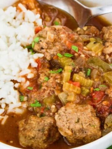 A white bowl contains white rice and a stew with meatballs, chopped celery, tomatoes, and sauce. A metal spoon rests in the bowl, and a green and white cloth is partly visible beside it.