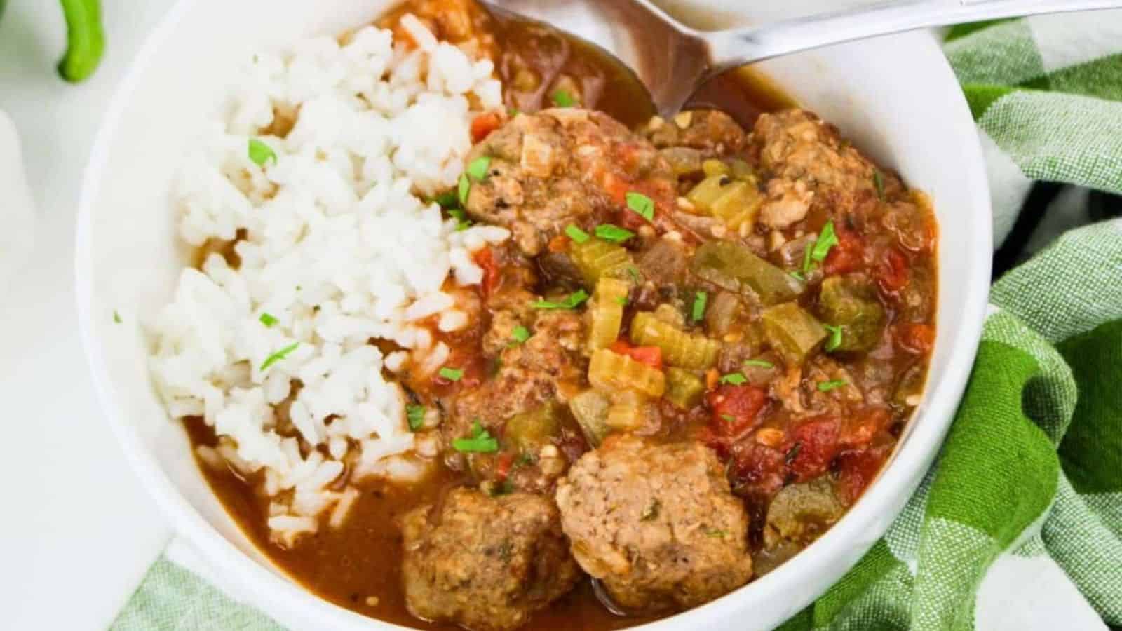 A white bowl contains white rice and a stew with meatballs, chopped celery, tomatoes, and sauce. A metal spoon rests in the bowl, and a green and white cloth is partly visible beside it.