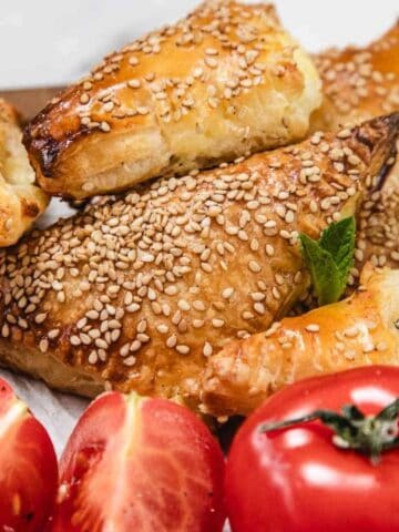 Golden brown sesame-covered pastry triangles are stacked on a plate alongside fresh mint leaves, halved red tomatoes, and a sliced hard-boiled egg. The pastries have a flaky, layered texture.