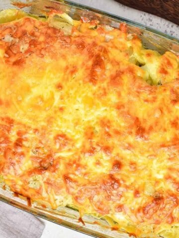 A rectangular glass baking dish filled with a baked casserole topped with melted, browned cheese sits on a white surface beside a folded gray cloth and a wooden utensil.