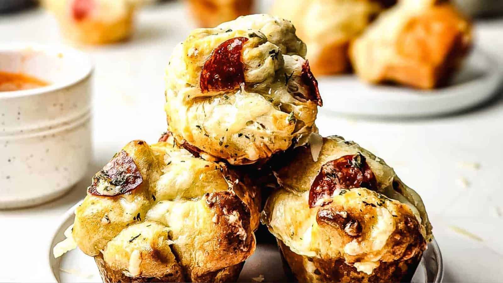 Three savory muffin-shaped pastries with melted cheese and slices of pepperoni on top are stacked on a plate. A cup of sauce and more pastries are visible in the blurred background.