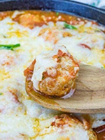 A wooden spoon holds a cheese-covered meatball over a cast iron skillet filled with meatballs in tomato sauce, topped with melted cheese and garnished with fresh basil leaves.